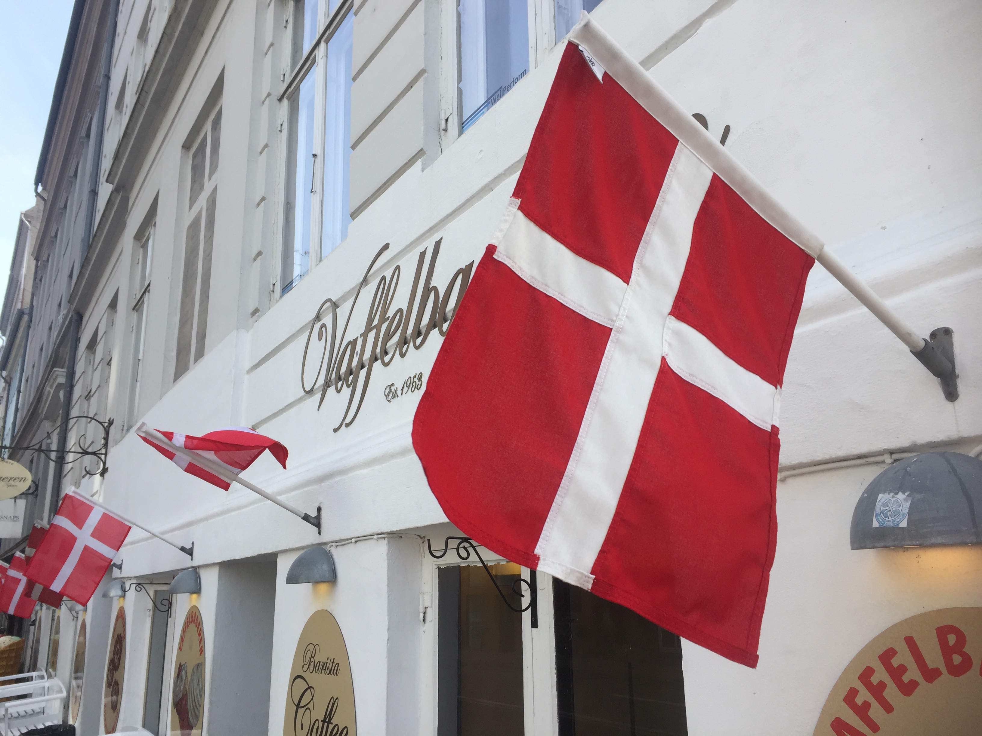Small Denmark flags hanging outside of white building. Red flag with off centered white cross.