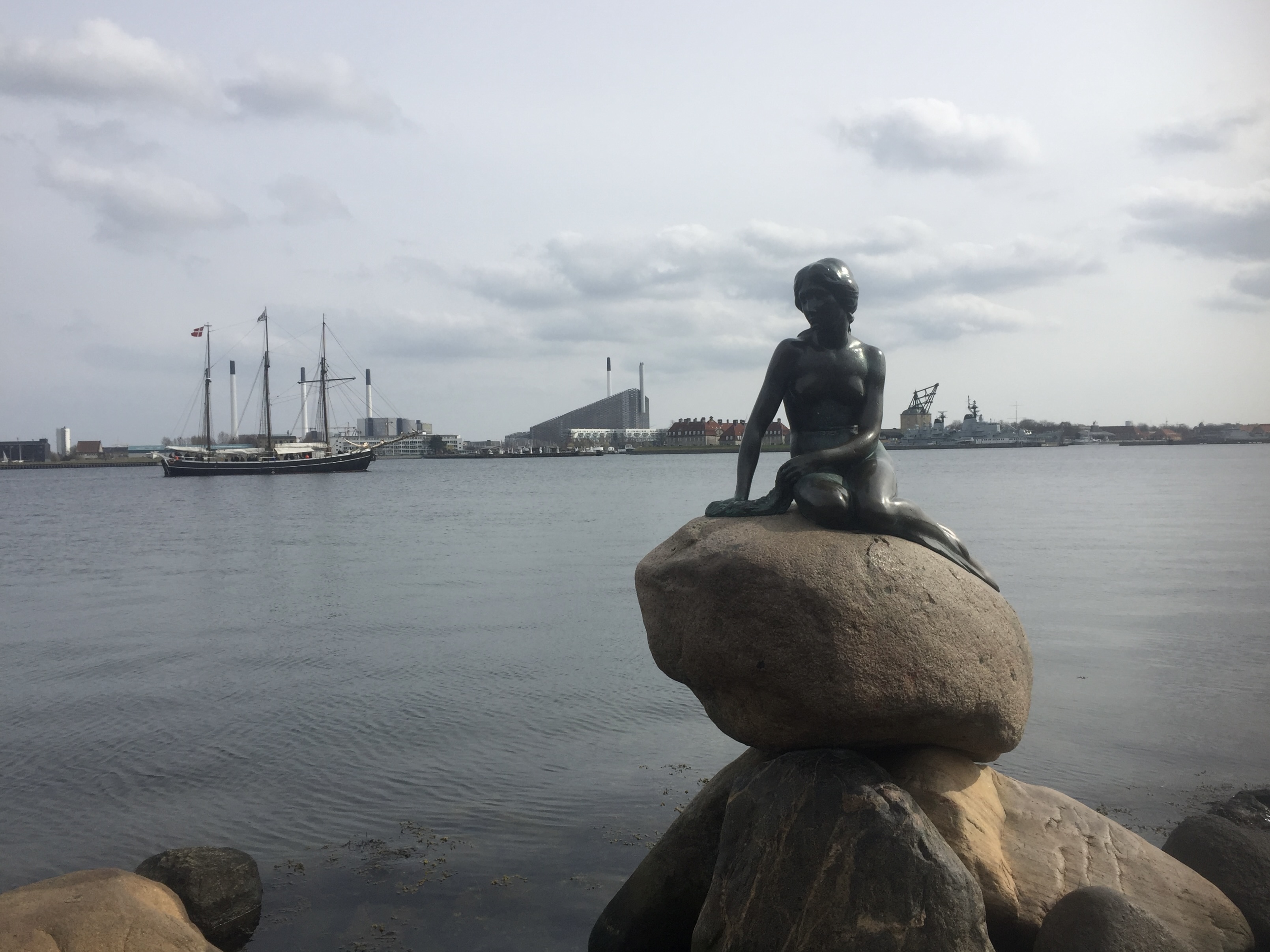 Little Mermaid statue sitting on a rock overlooking the ocean. Sail boat and land in the background.