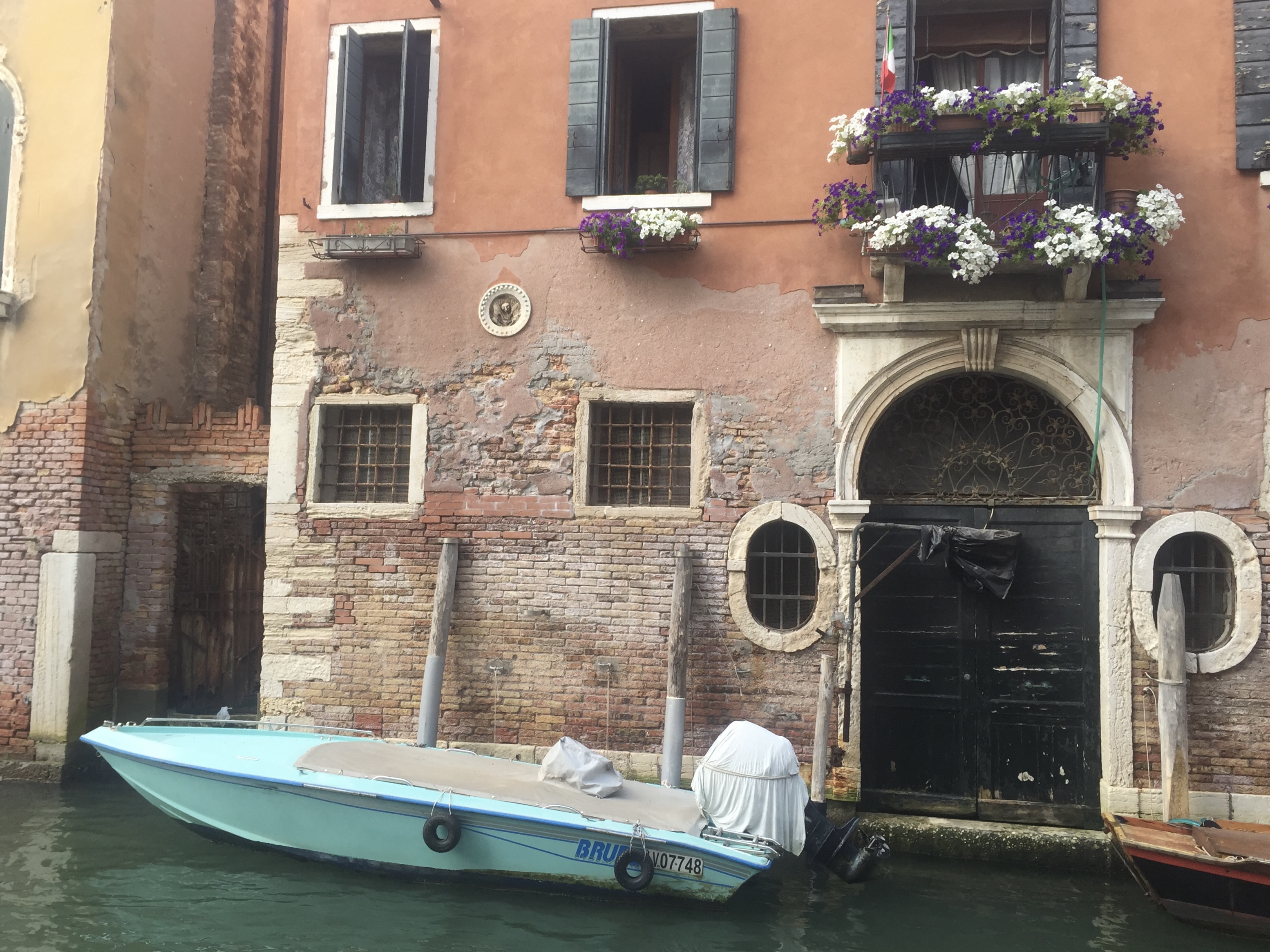 Tattered brick building with white and purple flowers along the canal of Venice. Small teal blue boat docked in front.